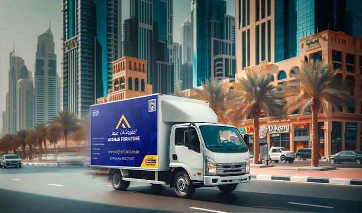 A white delivery truck with a blue "Asghar Furniture" logo drives past modern buildings in Dubai.