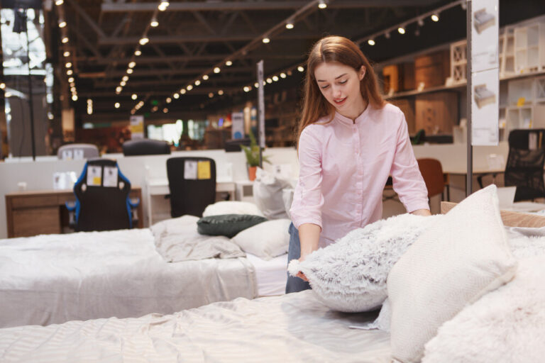 Young woman neatly arranging pillows on a comfortable bed display in a brightly lit furniture showroom.