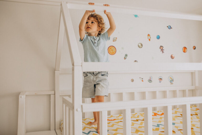 A playful child with curly hair standing in a white house-shaped bed, surrounded by space-themed wall decals.