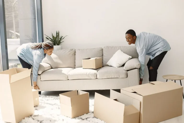 Two people in light blue shirts carefully moving a beige sofa in a room with cardboard boxes.