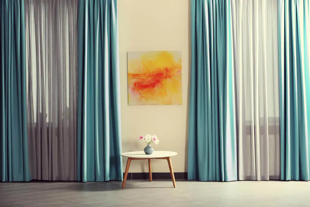 Modern interior with teal and sheer curtains, round wooden table, and abstract wall art.