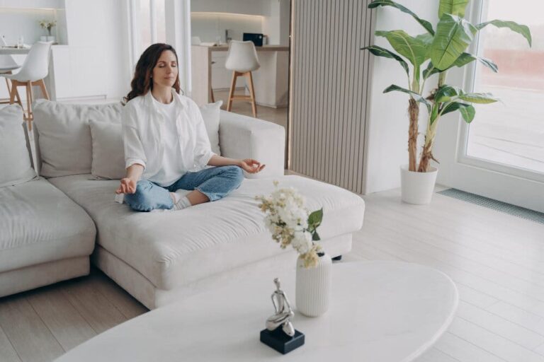 A woman meditates peacefully on a modern, light-colored sectional sofa in a bright and serene living room.