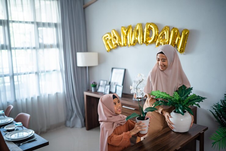 Two women, one adult and one child in hijabs, arranging plants with a 'RAMADAN' balloon decoration.