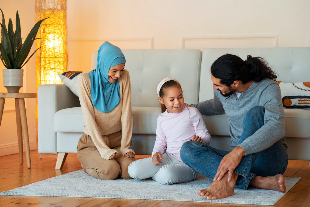 A happy family, including a woman in hijab, man, and girl, sitting playfully on a rug in a cozy home.