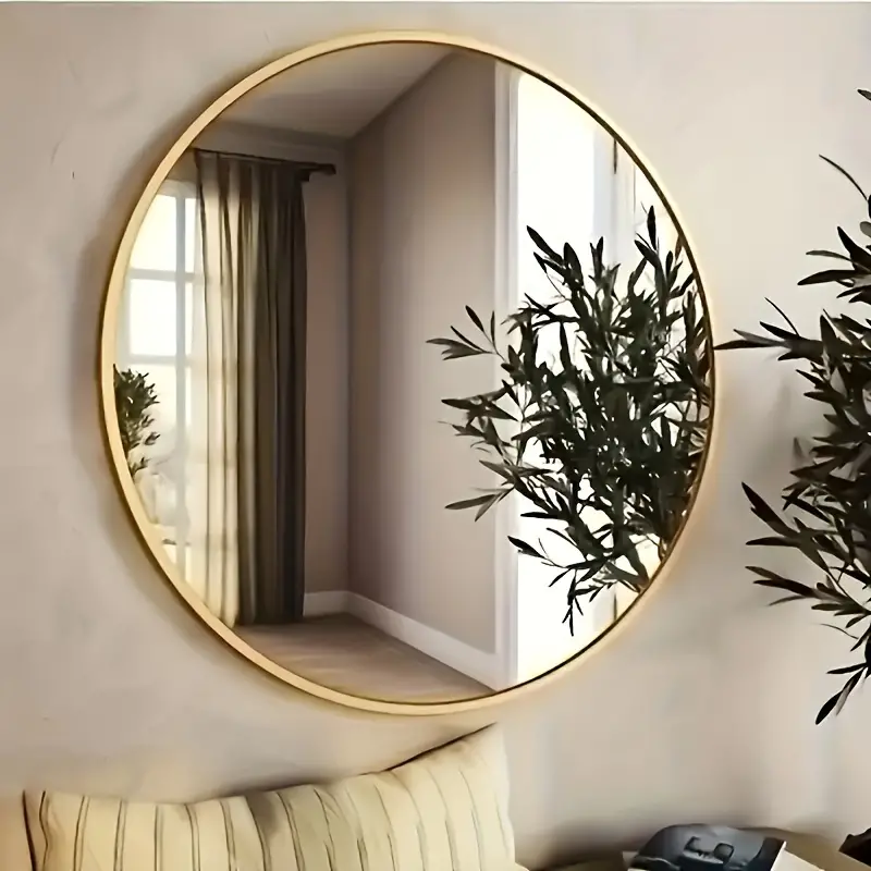 A round gold-framed mirror reflecting a window and a green plant, adding elegance to a room.